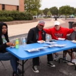 Four people sitting at a blue table outside pose for a photo at "A Pint For Kim" blood drive.