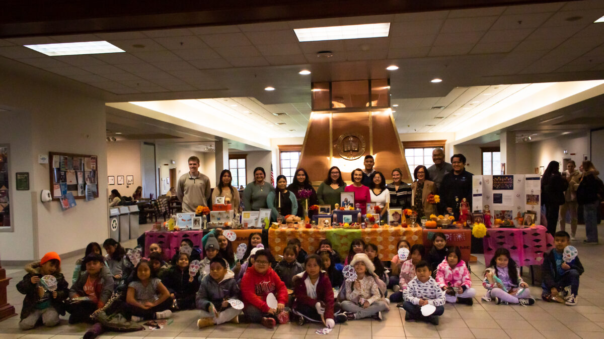 Elementary school students and Alpha Mu Gamma members pose by a colorful ofrenda (altar) in the Founders Lounge at Elmhurst University.