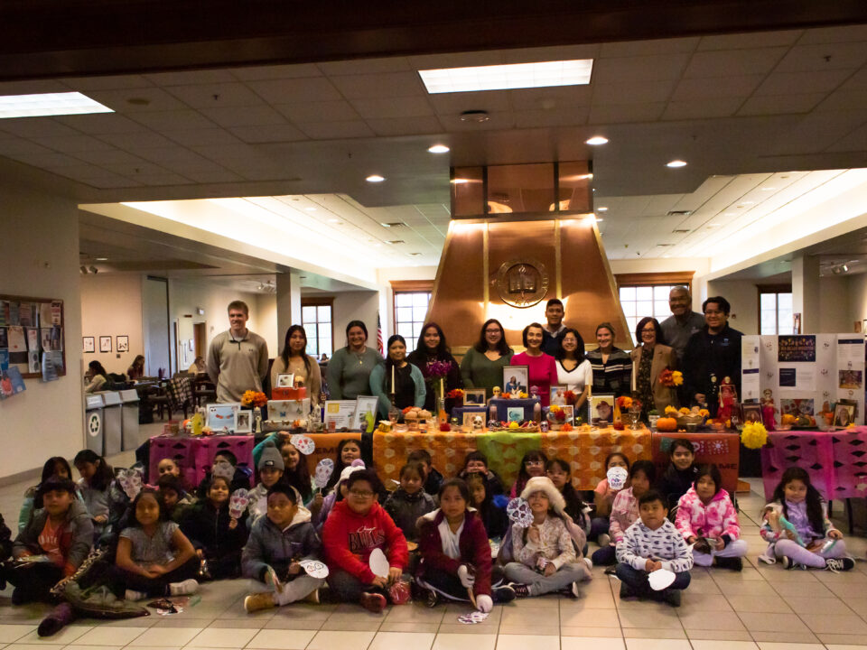 Elementary school students and Alpha Mu Gamma members pose by a colorful ofrenda (altar) in the Founders Lounge at Elmhurst University.